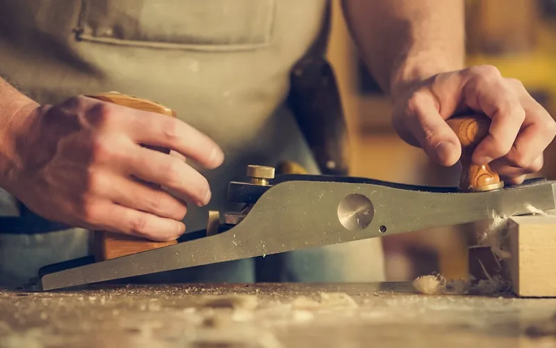 carpenter's hands using a hand plane to smooth a wood surface before finishing, with wood shavings