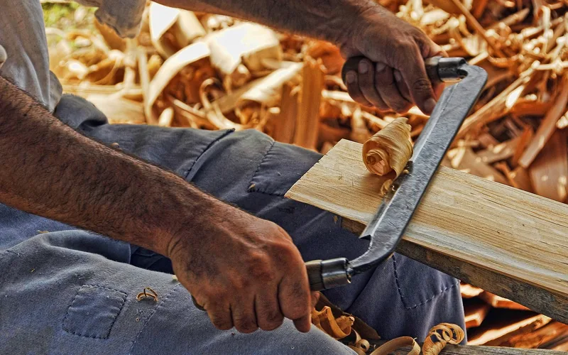 carpenter shaping a wood plank with a drawknife, wood shavings curling off the surface