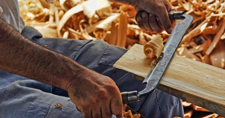 Carpenter shaving a wood plank by hand with a drawknife to shape a shelf bracket