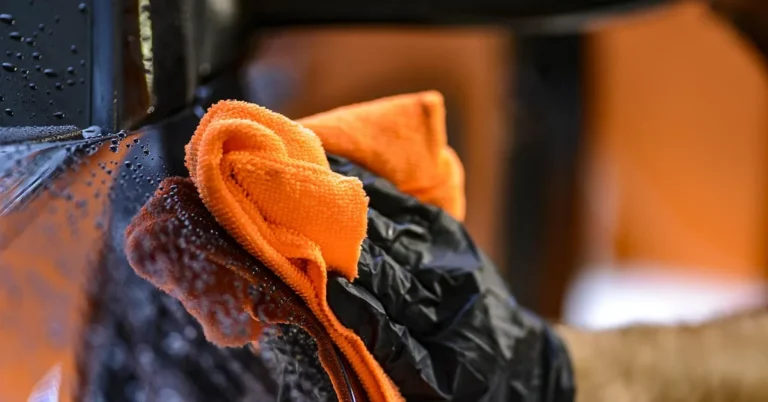 gloved hand wiping a car surface clean with an orange microfiber cloth
