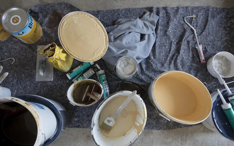 Open paint buckets, brushes, and a putty knife laid out on a drop cloth for a home paint job