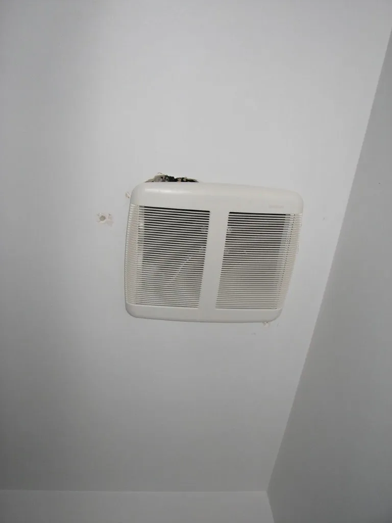 Close-up of a bathroom fan grille being measured with a tape measure near the ceiling
