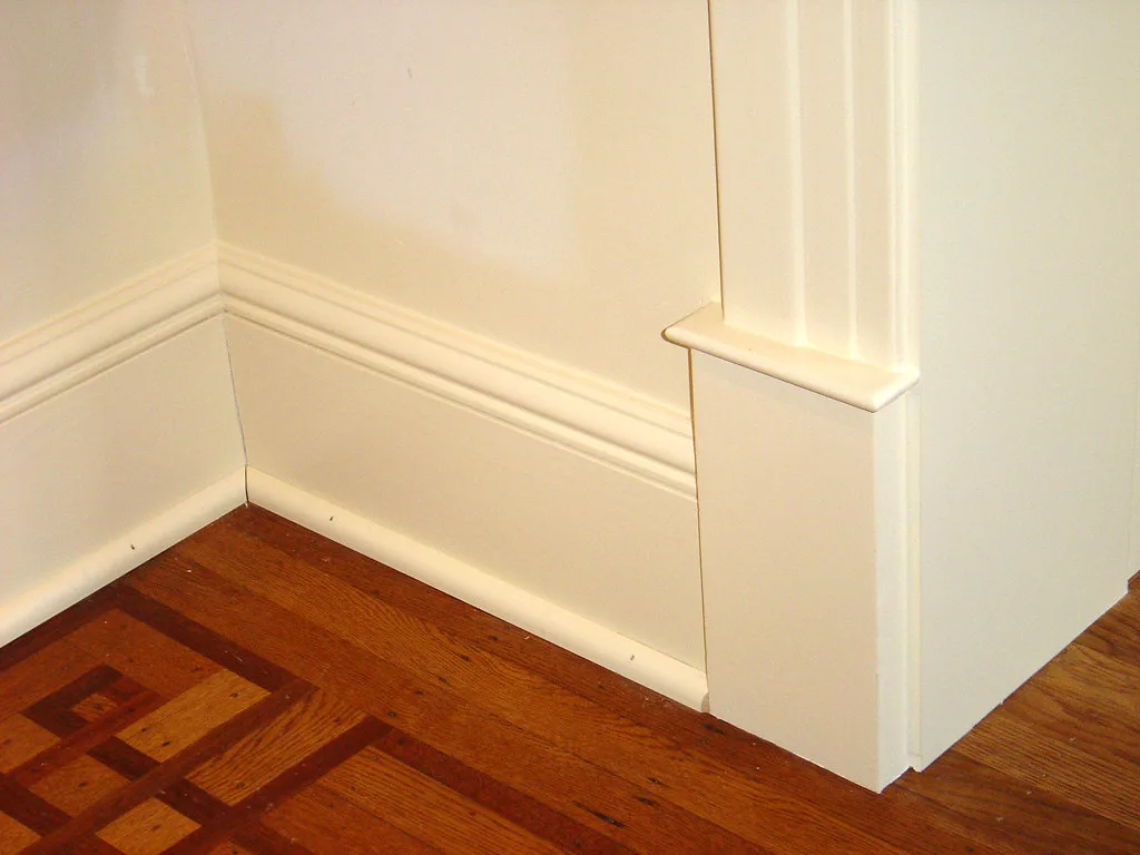 Homeowner compares caulk tubes beside painted baseboards and crown molding in a bright room
