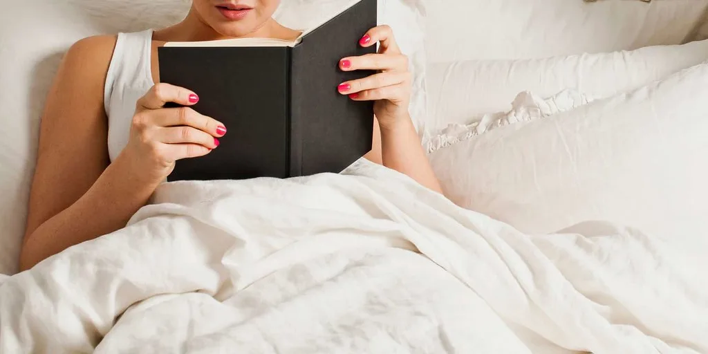A person on a bed holding their neck beside a mattress and pillow in natural light.