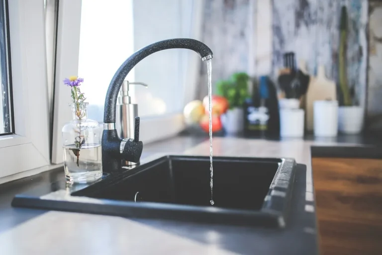 Water flows from a black faucet into a dark kitchen sink beside a glass vase with a purple flower on a countertop