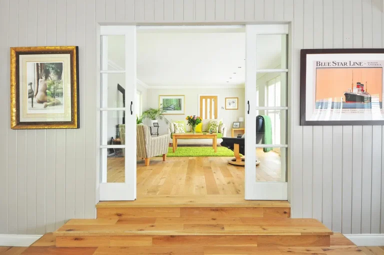 Open French doors reveal a bright living room with a sofa, coffee table, rug, and framed wall art