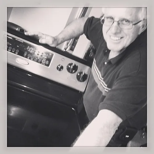 A smiling man stands beside a kitchen oven and touches its control panel