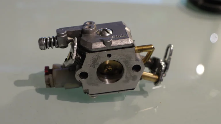 A silver Zama carburetor with brass fittings rests on a reflective white surface