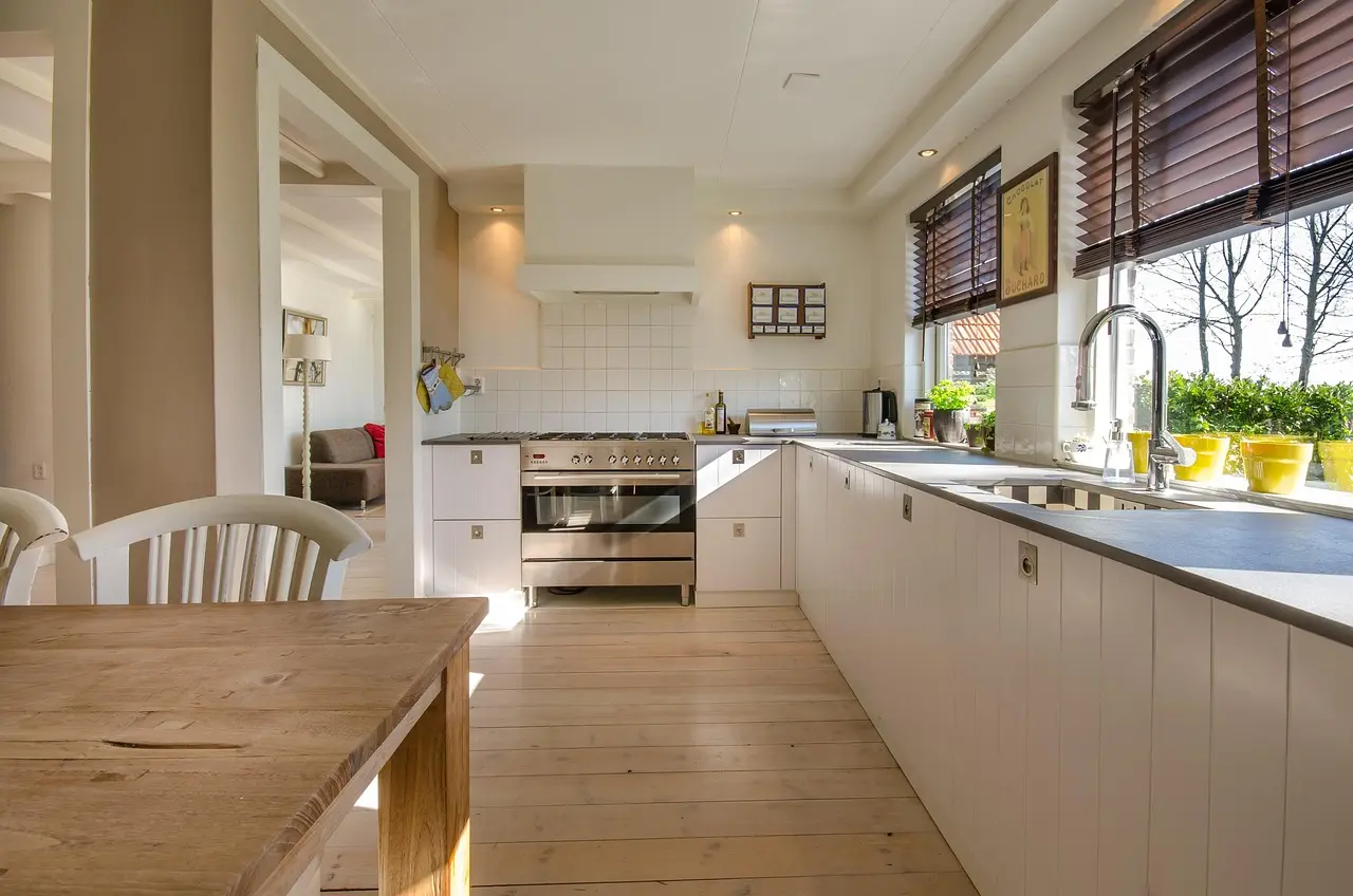 Bright kitchen with white cabinets, wood floor, steel stove, sink by window with blinds, and a wooden table in foreground