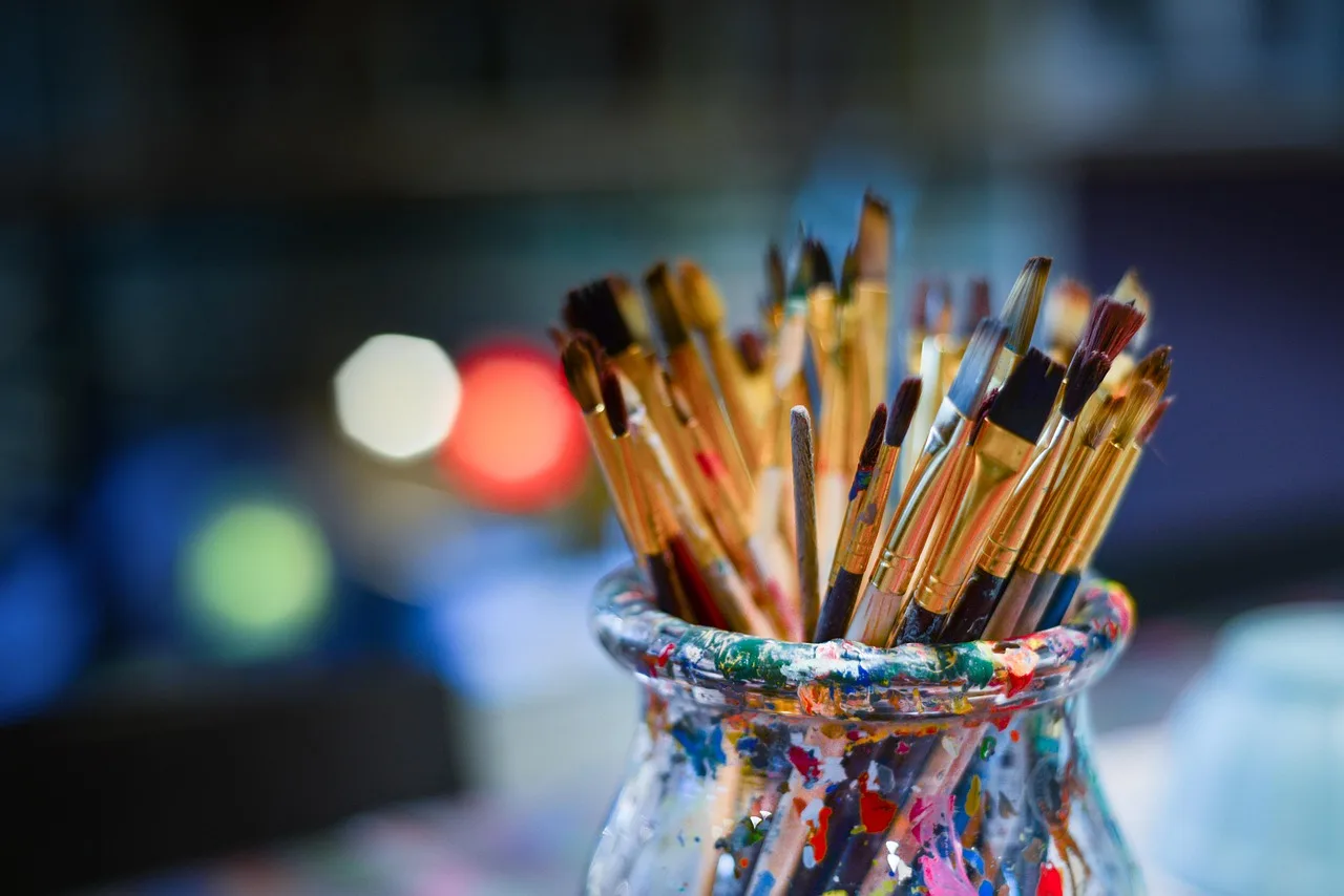 Paintbrushes stand in a paint-splattered glass jar against a blurred background with colorful bokeh lights