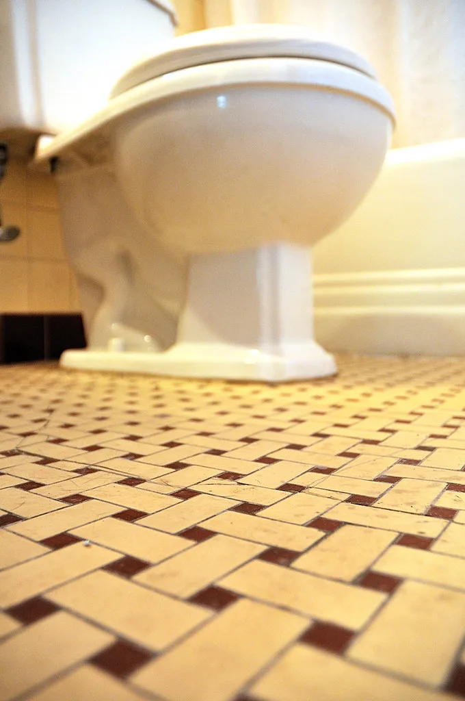 A hand wipes the toilet seat hinge and rim with a microfiber cloth beside a cleaner bottle.