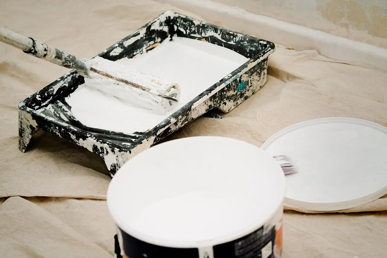 A paint roller rests in a white-filled tray beside an open paint can on a drop cloth